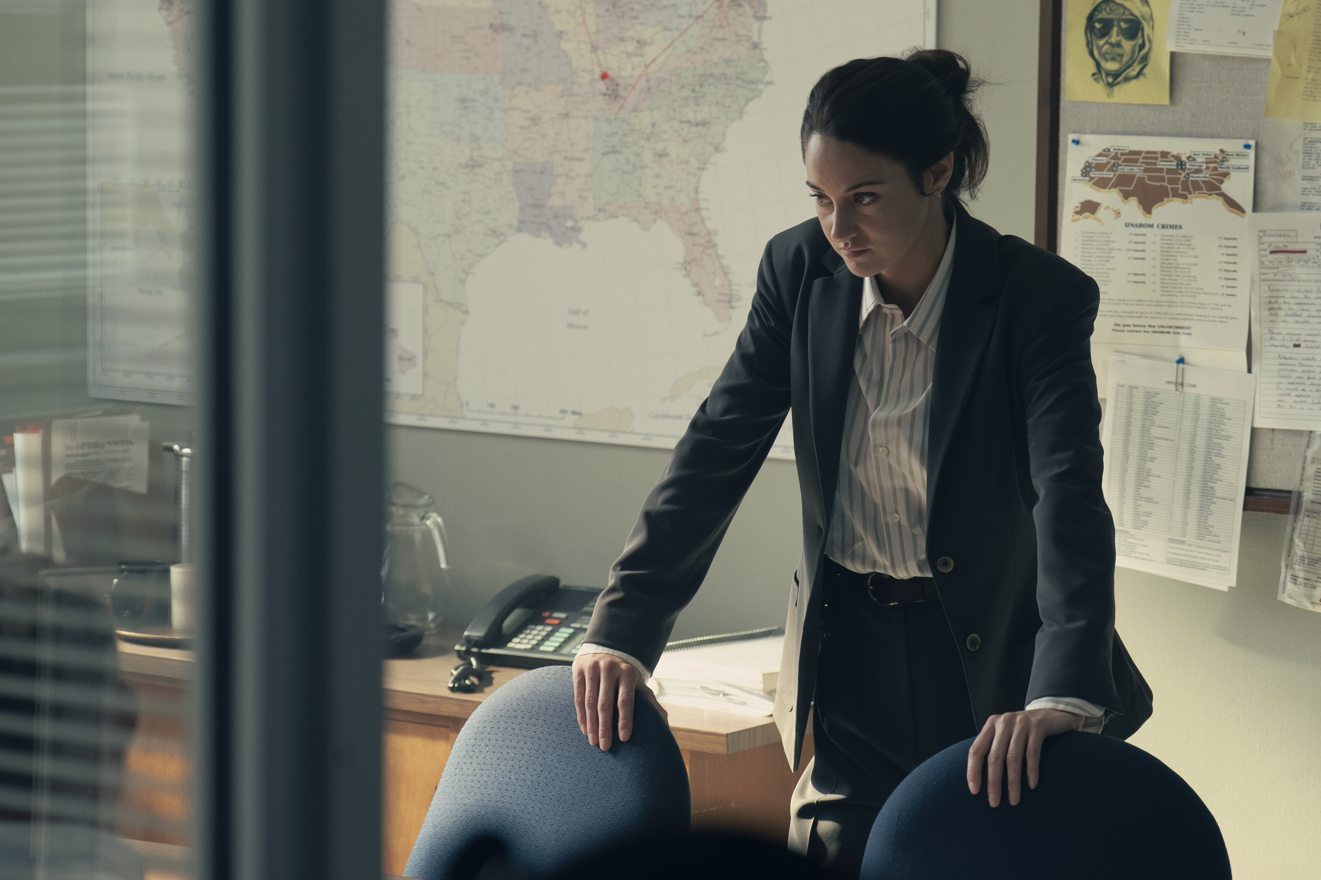 "A woman in a suit leans on a conference table in an office, appearing serious and focused. The background features a large map, bulletin board with notes, a desk phone, and documents, suggesting an investigative or professional setting."