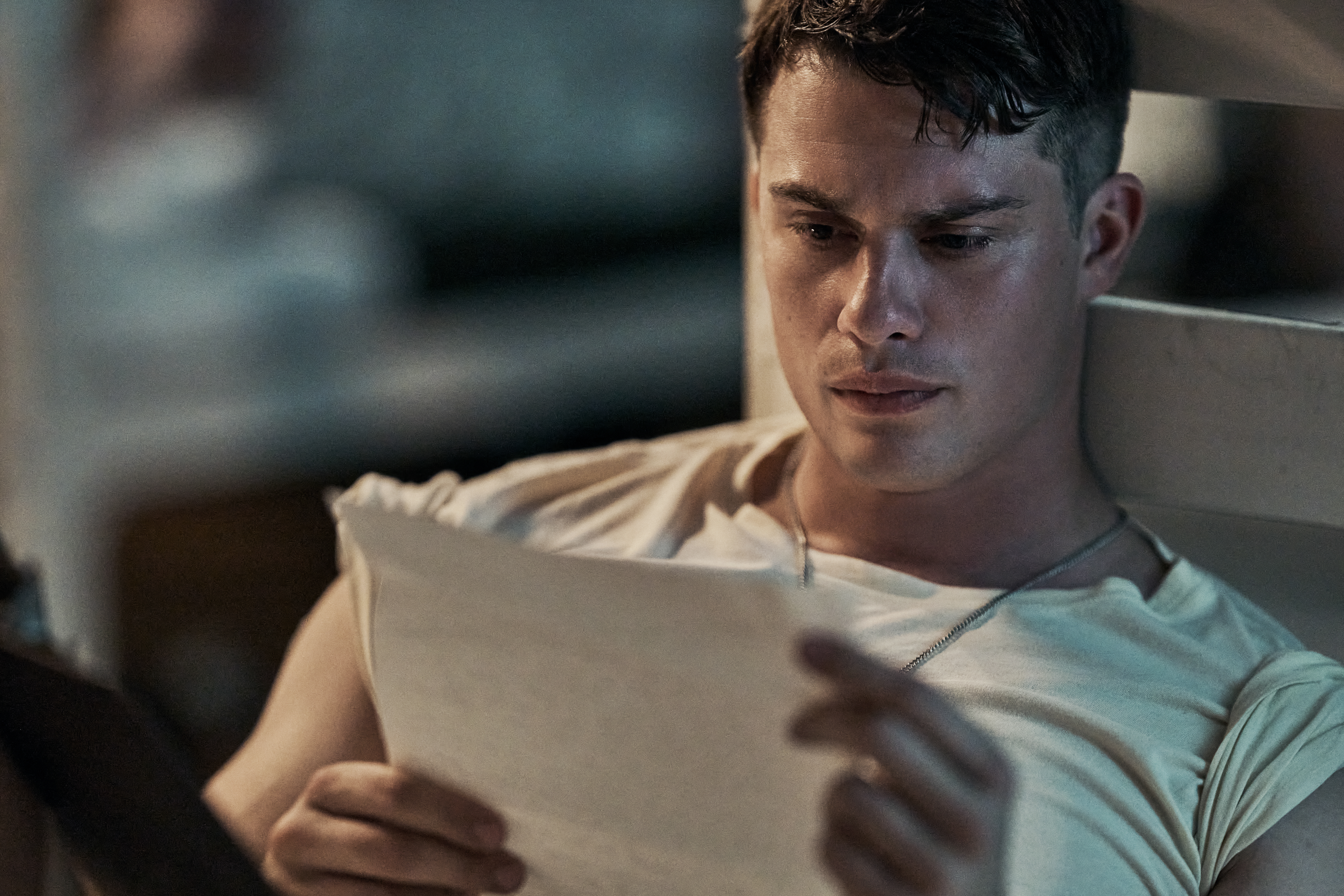 "Nicholas Galitzine as John McLaughry in a casual t-shirt sitting indoors, leaning against a wall or bed frame, reading a letter with a serious expression, background softly blurred, conveying a contemplative or emotional moment."