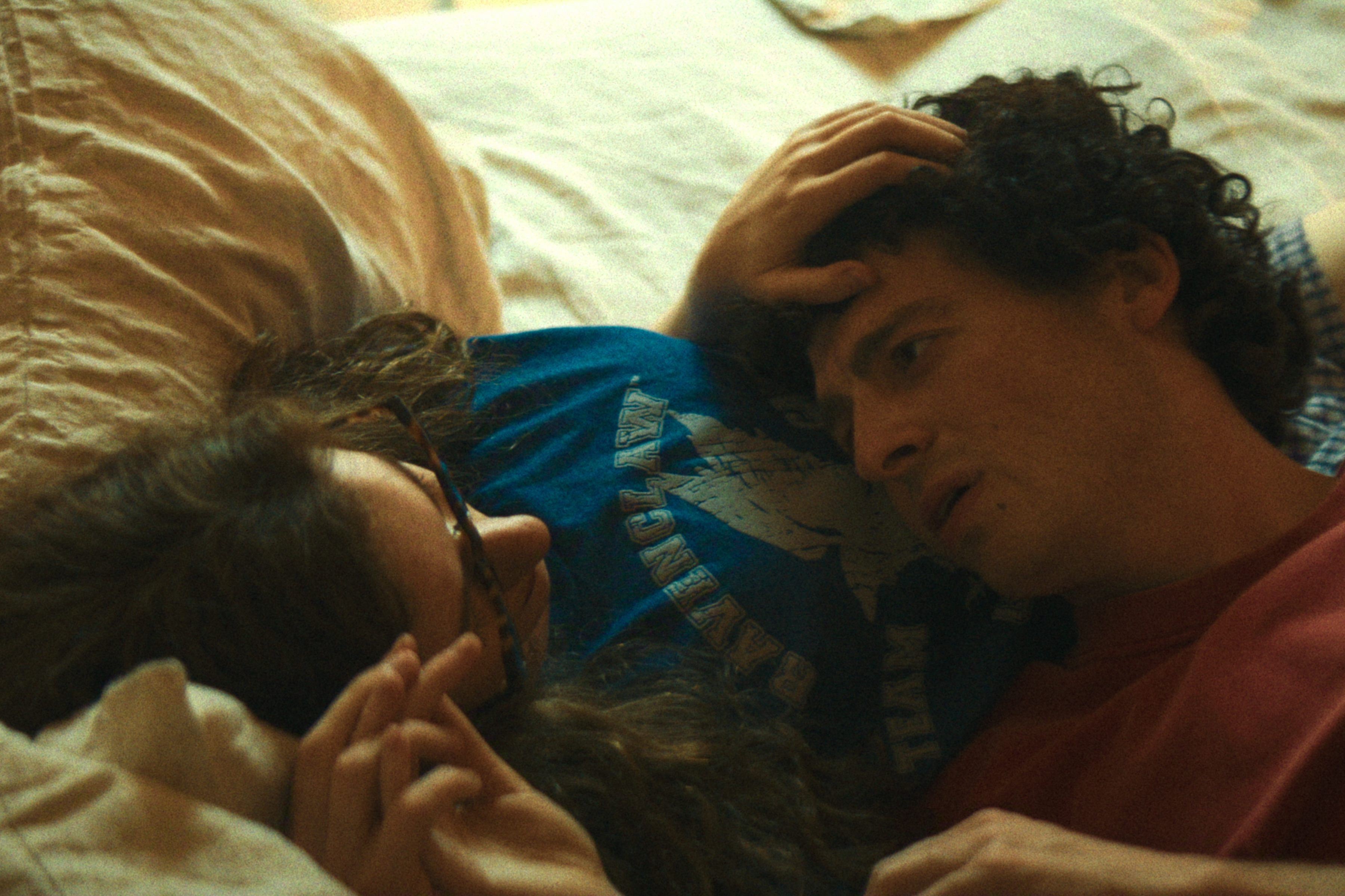 "Two people lying closely together on a bed in a cozy, dimly lit room, gazing at each other and sharing an intimate moment, creating a sense of warmth and connection."