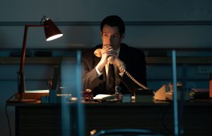 "A man in a suit sits alone at a desk in a dimly lit office, holding a telephone receiver, with a red desk lamp providing focused light on the work surface."