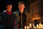 "Two young men stand together at a nighttime candlelight vigil, looking somber. One has his arm around the other for comfort. The setting is outdoors in an urban area, with lit candles in the foreground."