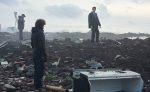 "Three people inspecting debris and rubble on a desolate, cloudy landscape; two stand among scattered wreckage and broken objects, while one observes from a distance, suggesting a post-disaster or crime scene investigation setting."