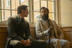 "Two men in period clothing sit on a wooden bench by a large window in a sunlit room, engaged in conversation. One wears a dark suit, the other a light suit and holds a cane, with brick buildings visible outside the window."