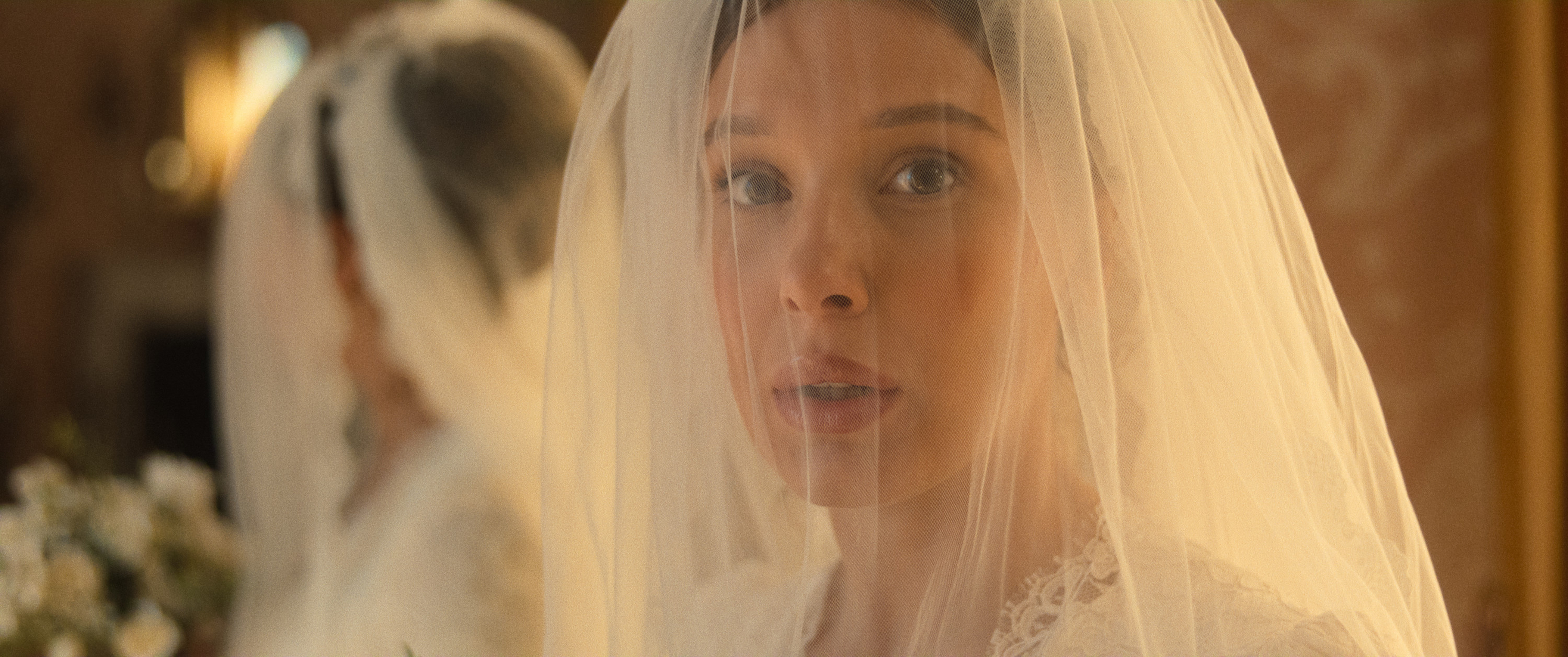 "A bride with a white veil and lace dress stands indoors, looking ahead with a soft expression. A mirror reflects her image, and flowers are visible in the background, creating an elegant, warm atmosphere."
