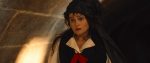 "Woman dressed in period costume with a black headdress and red ribbon stands in a dimly lit stone hallway, looking surprised or concerned. Warm light highlights the stone wall in the background."