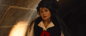 "Woman dressed in period costume with a black headdress and red ribbon stands in a dimly lit stone hallway, looking surprised or concerned. Warm light highlights the stone wall in the background."