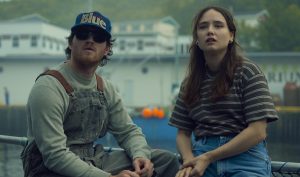 "Two people sit outdoors near a harbor, with fishing docks and buildings in the background. The man wears sunglasses, overalls, and a cap, while the woman wears a striped shirt and jeans. Both appear to be looking at something off-camera."