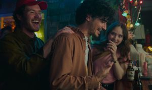 "Three friends laughing and talking at a bar with drinks, colorful lights, and a casual, lively atmosphere in the background."
