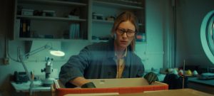 "A woman wearing glasses and gloves works in a laboratory, reaching into a container on a counter, surrounded by scientific equipment and shelves in a well-lit room."