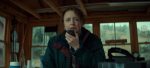 "A woman in a green jacket speaks into a handheld radio inside a wooden cabin with large windows, surrounded by communication equipment and natural outdoor light."
