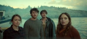 "Four people stand near a waterfront, looking concerned. Behind them is a boat and a rocky shoreline with pine trees under a cloudy sky. The lighting is moody, suggesting a dramatic or tense atmosphere."