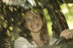 "Smiling woman sitting in driver’s seat of a car, seen through windshield with green leafy reflections, natural outdoor lighting, relaxed atmosphere."
