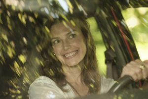 "Smiling woman sitting in driver’s seat of a car, seen through windshield with green leafy reflections, natural outdoor lighting, relaxed atmosphere."