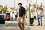 "Young man in black shirt and khaki shorts walking on a marina dock, with boats, flowers, and people in the background on a sunny day."