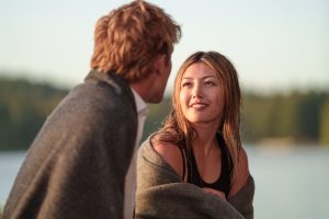 "Two people wrapped in a blanket sit by a calm lake at sunset, conversing and smiling, with trees in the background and soft natural lighting."