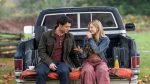 "Man and woman sitting on tailgate of a pickup truck in a grassy outdoor setting, talking and eating together, with drinks and food on a blanket, autumn trees in the background."