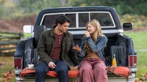 "Man and woman sitting on tailgate of a pickup truck in a grassy outdoor setting, talking and eating together, with drinks and food on a blanket, autumn trees in the background."
