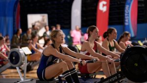 "Women are rowing at an indoor rowing race at a gym or sports event, focused and determined, with banners and spectators in the background."
