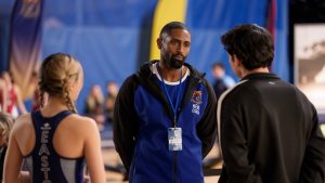 "A coach in a blue and black jacket stands in a gymnasium talking to two athletes, one male and one female, with sports banners and people blurred in the background."