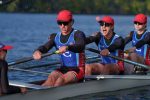 "Four male rowers in matching uniforms and red hats are rowing on a calm river in a race, with trees and clear sky in the background, showing focused teamwork and athletic effort."