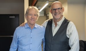 "Two smiling men stand side by side in an indoor office hallway, one with a hand on the other's shoulder. Both are dressed in business casual attire, with neutral-colored walls and a door in the background."