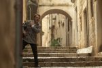 "Man in grey plaid blazer stands on stone steps in a narrow, historic European alleyway with archways and old buildings, appearing alert or cautious as he looks around the corner."