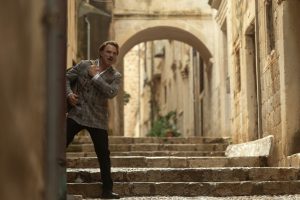 "Man in grey plaid blazer stands on stone steps in a narrow, historic European alleyway with archways and old buildings, appearing alert or cautious as he looks around the corner."