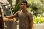 "Young man wearing a backwards cap and t-shirt stands outside beside an old, weathered van in a green, sunlit, outdoor setting with trees and a white fence in the background, looking ahead with a serious expression."