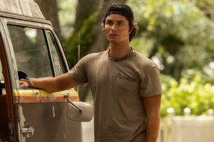 "Young man wearing a backwards cap and t-shirt stands outside beside an old, weathered van in a green, sunlit, outdoor setting with trees and a white fence in the background, looking ahead with a serious expression."