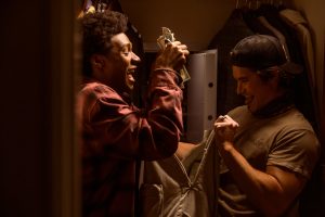 "Two young men excitedly hold cash in a dimly lit closet, with clothes hanging in the background and an open safe visible behind them, suggesting they just found or stole the money."