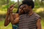 "Two people stand closely together outdoors. The woman holds up a knife while the man gently guides her hand. They both appear calm and focused, surrounded by lush greenery in a natural setting."
