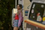 "Young woman with long hair wearing a red shirt and denim shorts leans against an RV in an outdoor wooded area, with another vehicle covered in stickers partially visible in the foreground."