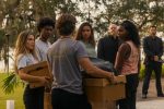 "A group of young adults stand outdoors holding cardboard boxes, facing each other with serious expressions; two men in black suits observe in the background near trees and grass."