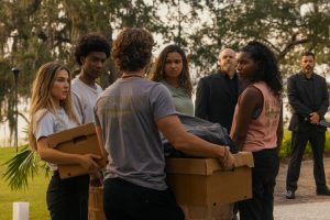 "A group of young adults stand outdoors holding cardboard boxes, facing each other with serious expressions; two men in black suits observe in the background near trees and grass."