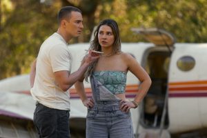 "Two people stand in front of a small plane in an outdoor setting, appearing to be in a serious conversation, with one gesturing while the other listens with arms on hips, surrounded by green trees."
