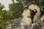"A person in mid-air falls or jumps on rocky terrain outdoors, surrounded by dust and trees, suggesting an action scene in a natural forest or mountain environment."