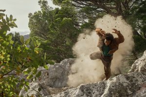 "A person in mid-air falls or jumps on rocky terrain outdoors, surrounded by dust and trees, suggesting an action scene in a natural forest or mountain environment."