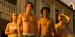 "Four young adults stand outside a large house in warm, golden sunlight. Two are shirtless, and the group looks intently ahead, suggesting curiosity or concern. The setting feels summery and relaxed."