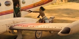 "Person riding a motorcycle reaches out to grab another person's hand from an open plane door as the plane takes off on a dirt runway, creating a dramatic action scene outdoors."