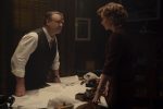 "A man and woman in formal attire have a serious discussion in a dimly lit office, standing over a desk covered with papers and old-fashioned equipment, creating a tense atmosphere."