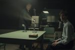 "Two men in formal attire are in a dimly lit interrogation room with a green table, microphones, and a film projector. One man stands by the projector while the other sits, suggesting a tense interview or investigation setting."