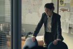 "A woman in a suit leans on a conference table in an office, appearing serious and focused. The background features a large map, bulletin board with notes, a desk phone, and documents, suggesting an investigative or professional setting."
