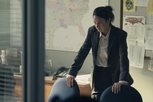 "A woman in a suit leans on a conference table in an office, appearing serious and focused. The background features a large map, bulletin board with notes, a desk phone, and documents, suggesting an investigative or professional setting."