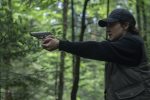"A person wearing a black cap and jacket holds a handgun and aims it in a dense, green forest, appearing alert and focused in a tense outdoor setting."