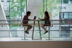 "Two people sit across from each other at a high table in a modern, glass-walled building, having a conversation. One uses a laptop, and greenery is visible outside the large windows."