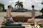 "Two people are talking on a sunny outdoor patio with a sofa, colorful pillows, and a table; palm trees and a large yacht are visible in the background, creating a relaxed, tropical setting."