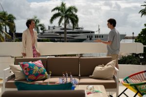 "Two people are talking on a sunny outdoor patio with a sofa, colorful pillows, and a table; palm trees and a large yacht are visible in the background, creating a relaxed, tropical setting."