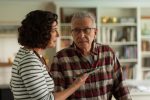 "A woman in a striped shirt talks to an older man in glasses and a plaid shirt in a home setting with bookshelves and decor in the background. The woman is holding a remote control toward the man."