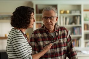 "A woman in a striped shirt talks to an older man in glasses and a plaid shirt in a home setting with bookshelves and decor in the background. The woman is holding a remote control toward the man."