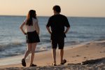"Two people walking along a sandy beach at sunset, facing the sea and each other, with gentle waves rolling in the background and soft evening sunlight illuminating the shoreline."